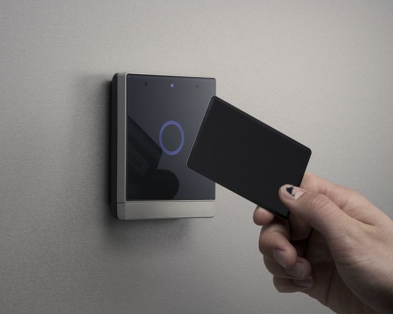 Hand presenting a credential card to a flush glass access reader on a grey wall